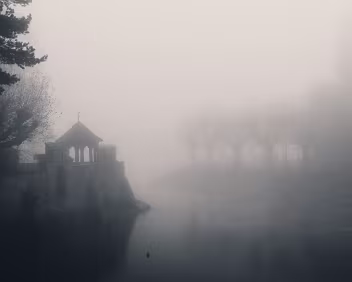 Fog Chapel Small stone riverside structure and nearby trees dissolve into dense mist over still water.
