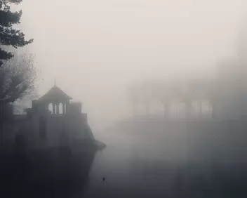 Fog Chapel Small stone riverside structure and nearby trees dissolve into dense mist over still water.